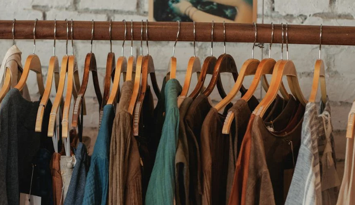 A wooden clothes rail holding a row of wooden hangers with assorted garments in earthy tones, including jackets, shirts and tops, set against a light brick wall background.